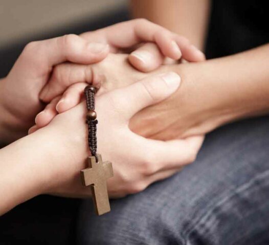 Två händer håller varandra med ett träkors i bakgrunden.